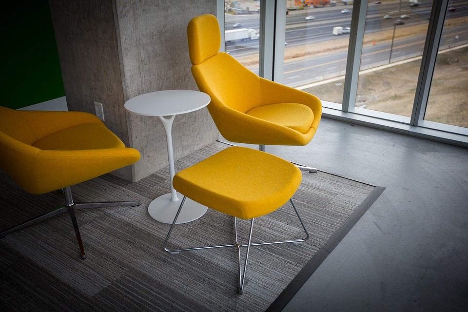 Modern lounge area with two yellow armchairs, a small white round table, and a matching ottoman on a gray rug.