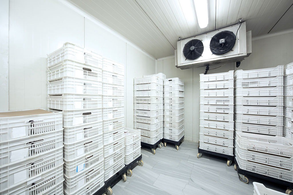 A clean, organised storage room with white plastic crates stacked on wheeled carts with a large vent fan overhead.