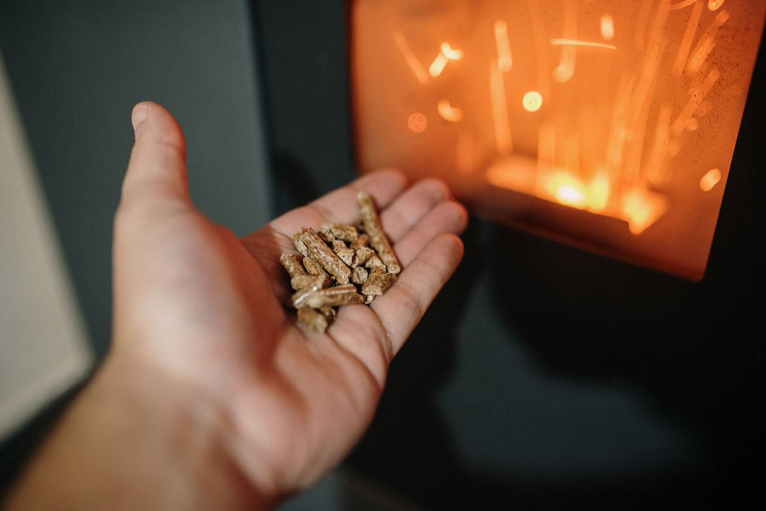 A hand holding small wood pellets near a glowing pellet stove, with visible sparks.