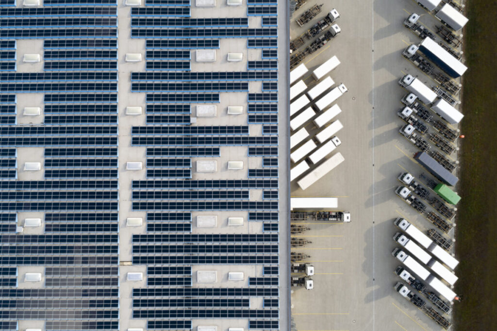 Aerial view of a large warehouse roof covered with solar panels, adjacent to a parking lot filled with rows of parked trucks and trailers.