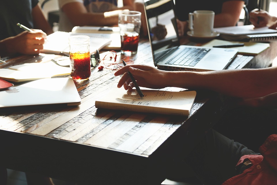 Hands writing in notebooks and typing on laptops at a wooden table, with glasses and coffee cups around.
