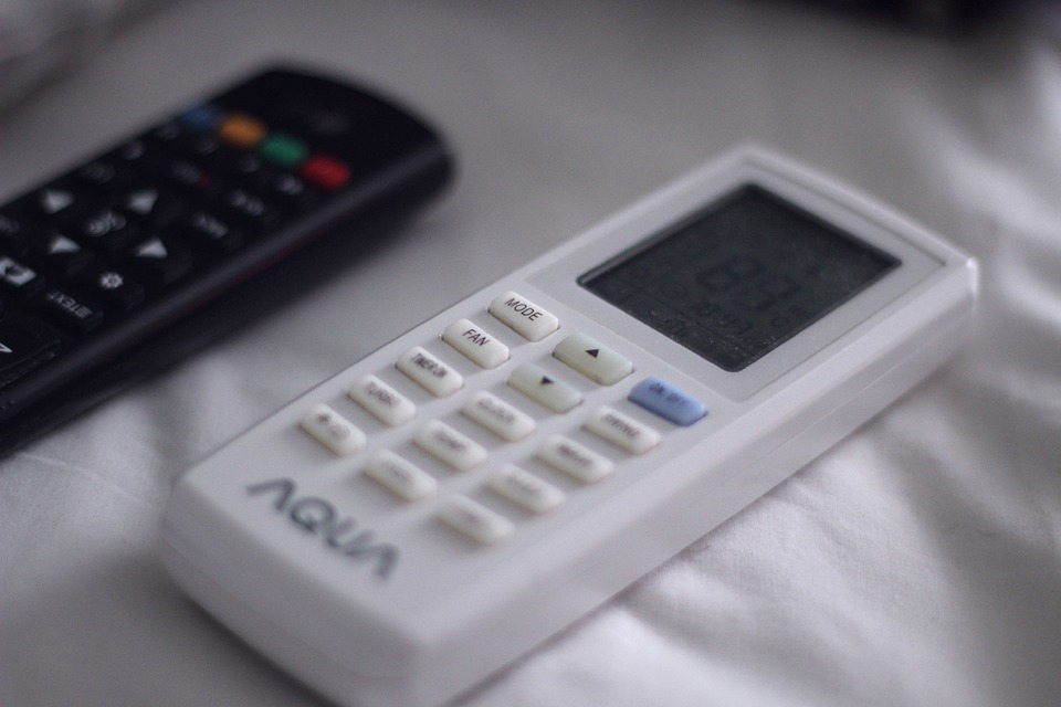 A white remote with soft buttons and a digital display rests on a white surface beside a black remote.