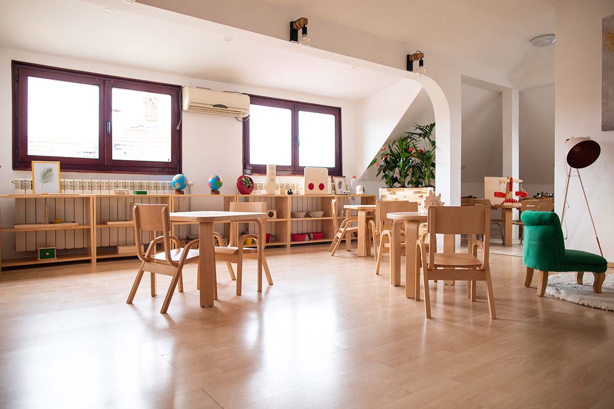 Classroom with wooden tables and chairs, shelves holding educational toys and materials, large windows, and a cozy green chair.