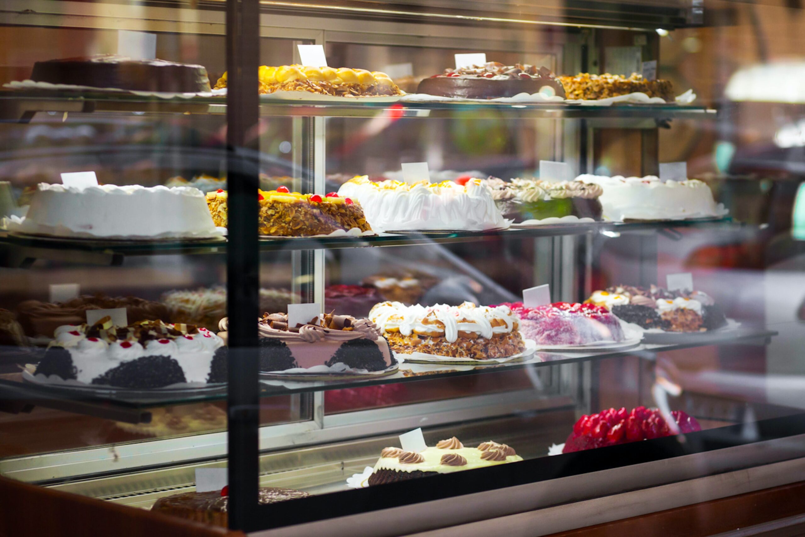 A display case with three shelves showcases a variety of colorful, elaborately decorated cakes.