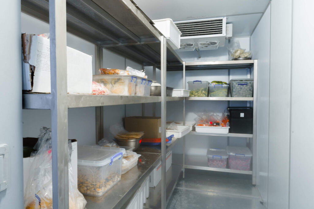 Commercial kitchen cold-room interior with metal shelves stacked with labeled plastic containers holding various frozen foods.