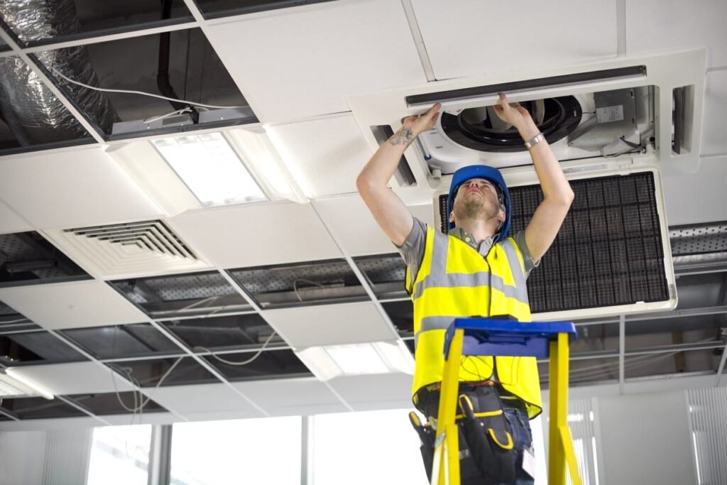 A construction worker in a high-visibility vest and hard hat stands on a ladder, repairing a ceiling air vent in an office.