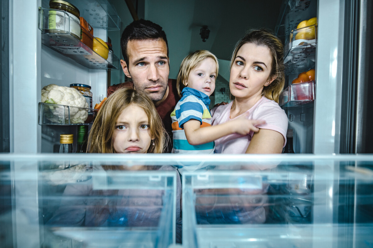 A family of four, peers into an open fridge looking concerned. The fridge shelves are nearly empty.