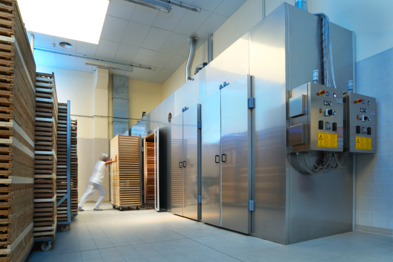 A worker in white pushes a wooden trolley towards a large, industrial stainless steel cold-room.